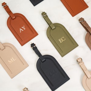 May include: Nine leather luggage tags with gold lettering. The tags are in various colors, including brown, beige, black, and green. The letters on the tags spell out the initials AY, ML, EC, JS, and GG.