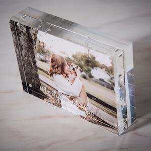 May include: A clear acrylic photo frame with a photo of a woman holding a dog. The photo is printed on the back of the frame and is visible through the clear acrylic. The frame is sitting on a white surface. The text "family forever" is engraved on the bottom of the frame.