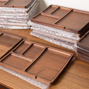 May include: Multiple stacks of wooden serving trays with a natural brown finish. Each tray has multiple compartments of varying sizes. The trays are on a wooden surface and wrapped in bubble wrap.