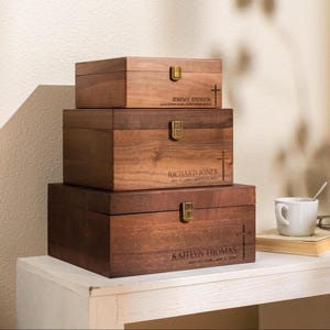 May include: Three wooden boxes stacked on top of each other. The boxes are a dark brown color with gold-colored clasps. Each box has a name and a cross symbol engraved on it. The boxes are sitting on a white table.