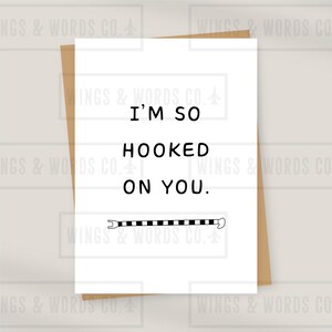 Può includere: Un biglietto di auguri bianco con la scritta "I'M SO HOOKED ON YOU" in grassetto, in nero. Sotto il testo c'è un'illustrazione di amo da pesca a strisce bianche e nere. Il biglietto è su una busta marrone.