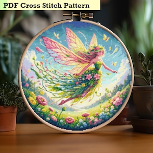 May include: A cross stitch pattern featuring a colorful fairy with pink wings and a green dress, surrounded by flowers and butterflies. The pattern is displayed in a wooden embroidery hoop.