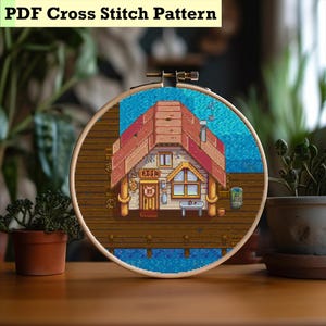 May include: A cross-stitch design in a wooden hoop. The image shows a pixelated cabin with a red roof, set against a blue water backdrop. The text "PDF Cross Stitch Pattern" is at the top. The cabin has a sign that says "FISH".