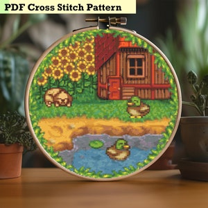 May include: A cross stitch pattern featuring a red and brown house with a green roof, surrounded by sunflowers and green grass. Two ducks are swimming in a blue pond with lily pads. The pattern is displayed on a round wooden hoop.