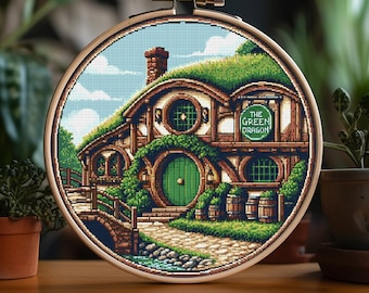 Puede incluir: Un patrón de punto de cruz de una casa hobbit verde, marrón y blanca con una puerta redonda verde y un letrero que dice "The Green Dragon".
