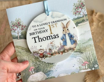 May include: A personalized 1st birthday ornament featuring Peter Rabbit and friends. The circular ornament has a light blue ribbon and reads "To a lovely grandson on your 1st birthday Thomas." The background is a watercolor scene with trees, a stream, and mushrooms.