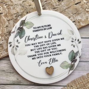 May include: A white round ornament with a green and white floral design. The ornament has a message that reads "To my future parents in law Christine & David You may not have given me the gift of life. But life has given me the gift of you. Thank you for welcoming me into your family. Love Ellie".