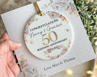 May include: A white card with a gold circle ornament featuring a floral design and the text "Congratulations Nanny & Grandad 50 Wedding Anniversary 1974-2024". The card also has the text "Love Alice & Thomas" at the bottom.