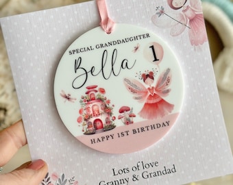 May include: A round white ornament with a pink ribbon, featuring a fairy illustration, a mushroom house, and the text: SPECIAL GRANDDAUGHTER, Bella, 1, HAPPY 1ST BIRTHDAY. It is attached to a square card with white polka dots and floral accents.