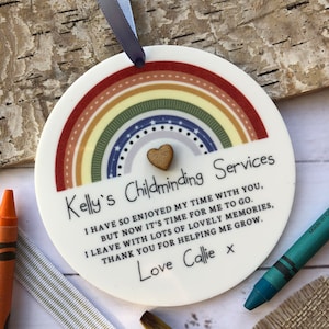 May include: A round, white ceramic ornament with a rainbow design and a small wooden heart in the centre. The ornament has the text "Kelly's Childminding Services" and a message of thanks from Callie.