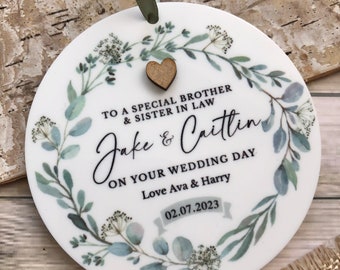 May include: White round ornament with a green leafy wreath design. The ornament has a wooden heart in the center and the text "To a special brother & sister in law Jake & Caitlin On your wedding day Love Ava & Harry 02.07.2023"