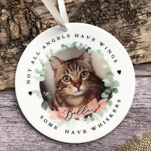 May include: A round white ceramic ornament featuring a photograph of a brown tabby cat. The text reads "NOT ALL ANGELS HAVE WINGS SOME HAVE WHISKERS" and "Bella" on a pink ribbon. It is decorated with green eucalyptus leaves and two small black hearts.