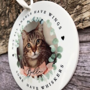 May include: A round white ceramic ornament featuring a photograph of a brown tabby cat. The text reads "NOT ALL ANGELS HAVE WINGS SOME HAVE WHISKERS" and "Bella" on a pink ribbon. A white ribbon loop is attached to the top for hanging.
