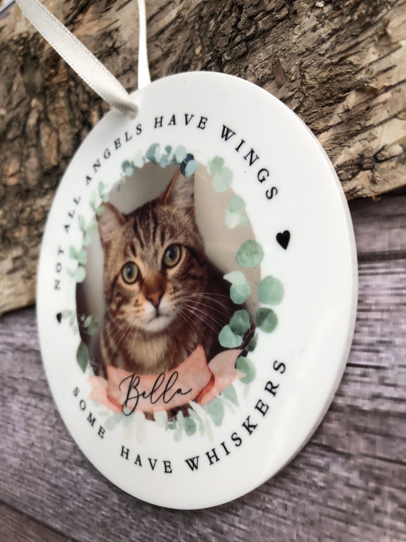 May include: A round white ceramic ornament featuring a photograph of a brown tabby cat. The text reads "NOT ALL ANGELS HAVE WINGS SOME HAVE WHISKERS" and "Bella" on a pink ribbon. A white ribbon loop is attached to the top for hanging.