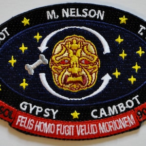 May include: A blue and red embroidered patch with a white oval containing a yellow and red face with a bone and arrows. The text "C.T. ROBOT M. NELSON T. SERVO GYPSY CAMBOT SOL FELIS HOMO FUGIT VELUD MORIONEM 903" is written around the oval.