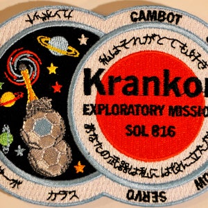 Puede incluir: Un parche circular blanco y azul con un círculo rojo en el centro. El círculo rojo tiene el texto "Krankor" en negro, "Exploratory Mission" en negro y "Sol 816" en negro. El círculo exterior tiene el texto "CamBot" en negro, "Gypsy" en negro, "Nelson" en negro y "Servo" en negro. El parche también tiene una imagen en blanco y negro de un planeta con un sistema de anillos y una estrella.