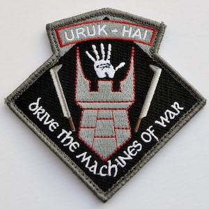 Pode incluir: Um remendo bordado preto e cinza com o texto "URUK-HAI" em vermelho e branco. O remendo apresenta uma torre cinza com uma impressão de mão branca no topo e duas espadas cinzas de cada lado. O texto "drive the machines of war" está escrito em um arco curvo ao redor da torre.