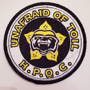 May include: A round, white and black embroidered patch with a yellow flower and a badger inside. The text "UNAFRID OF TOIL H.P.Q.C." is around the edge.