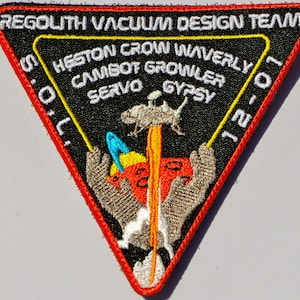 May include: A triangular patch with a red border and black background. The patch features a white embroidered design of a robotic arm holding a red sphere with a yellow flame coming out of it. The text "REGOLITH VACUUM DESIGN TEAM" is embroidered in yellow at the top of the patch. The text "HESTON CROW WAVERLY", "CAMBOT GROWLER", "SERVO GYPSY", "5.O,L.", and "12-01" are embroidered in white around the design.