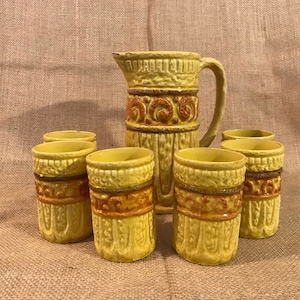 May include: A set of yellow ceramic pitcher and six cups with brown geometric designs. The pitcher has a handle and a spout. The cups are all the same size and have a rounded shape.