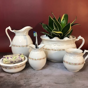 May include: A set of five white ceramic bathroom accessories with floral designs. The set includes a pitcher, a soap dish, a toothbrush holder, a small pitcher, and a mug.