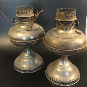 May include: Two antique silver oil lamps with ornate metal tops and bases. The lamps have a distressed finish and are in good condition.
