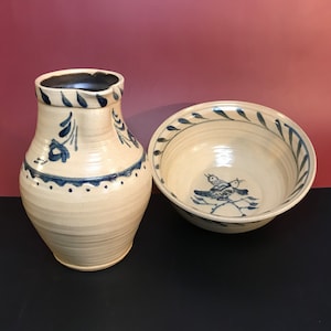 May include: A set of two handmade ceramic pieces, a pitcher and a bowl, both with a cream-colored glaze and blue floral designs. The pitcher has a rounded body and a spout, while the bowl has a wide rim and a bird design.