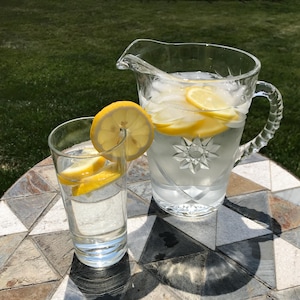 May include: A clear glass pitcher and a glass of lemonade with lemon slices. The pitcher is filled with ice and lemon slices. The glass of lemonade has a lemon slice on the rim. The pitcher and glass are on a stone table.