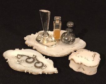 May include: A white porcelain vanity set with a tray, a small box, and a dish. The tray has a crystal bottle stopper, a clear glass bottle with a gold cap, and a silver bottle with a silver stopper. The dish has a pair of silver earrings.