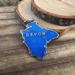 Pode incluir: Um amuleto de prata em forma do condado de Devon, Inglaterra. O amuleto é azul com contorno prateado e a palavra "DEVON" em letras prateadas.