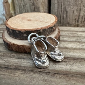 Könnte beinhalten: Ein silberfarbener Metallanhänger in Form eines Paares kleiner Babyschuhe, die durch einen einzelnen Ring verbunden sind. Jeder Schuh hat eine abgerundete Spitze, einen kleinen Zierknopf an der Vorderseite und ein hohles Inneres mit Metalloberfläche.