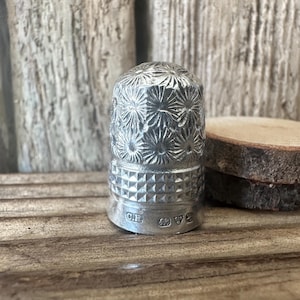 Puede incluir: Un dedal de plata con un diseño floral y una marca de fabricante en la parte inferior. El dedal está sobre una superficie de madera.