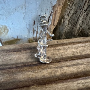 May include: A silver-toned charm depicting a person operating a water pump. The charm has a loop at the top for attachment and is detailed with the pump's handle and base. The charm is set against a rustic wooden background.