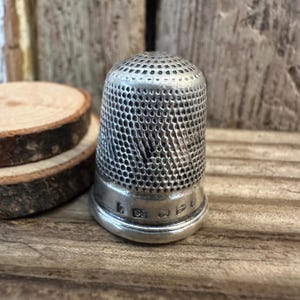 Puede incluir: Un dedal de plata con una superficie texturizada y un patrón. El dedal tiene un borde elevado y una pequeña inscripción en la parte inferior.