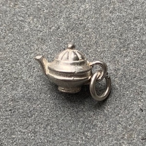 Könnte beinhalten: Ein silberner Anhänger in Form einer Teekanne. Die Teekanne hat einen runden Körper und einen Deckel mit einem Knopf oben. Der Anhänger hat eine Schlaufe zum Anbringen an einem Armband oder einer Halskette.