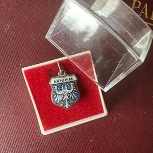 Op de afbeelding: Een zilveren bedel met een blauw geëmailleerd schild met een witte adelaar met een rode kroon en het woord "ARNHEM" in witte letters. De bedel zit in een rood fluwelen doosje met een witte rand.