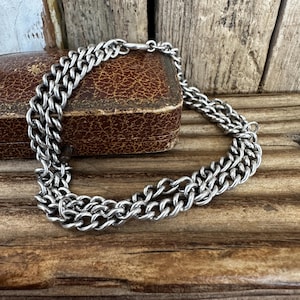 Op de afbeelding: Een zilverkleurige metalen kettingarmband met een dubbellaags stoeprandontwerp. De armband is geconstrueerd met in elkaar grijpende ovale schakels die een continu, uniform patroon door het hele stuk creëren, afgewerkt met een eenvoudige metalen sluiting.