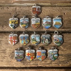 May include: A collection of vintage shield-shaped pendants, each featuring a different city or region. The pendants are silver-toned with colorful enamel designs, including cityscapes, flags, and coats of arms. The word "ESPAÑA" is visible on one pendant.