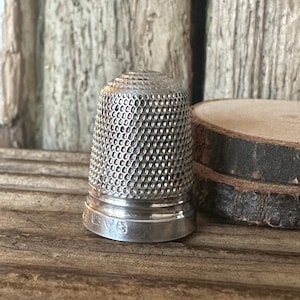 Puede incluir: Un dedal de plata con una superficie texturizada y una marca de contraste en la parte inferior.