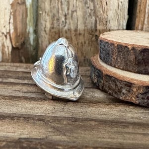 May include: A small, silver-colored miniature firefighter's helmet. The helmet has a detailed design with a crest and a brim. The background includes wooden elements, suggesting a rustic or handcrafted setting.