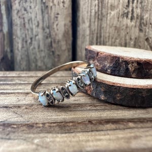 Könnte beinhalten: Ein goldfarbener Ring mit einer Reihe ovaler Opale. Jeder Opal ist in einer silberfarbenen Fassung gefasst, was einen Kontrast zum goldenen Band erzeugt. Der Ring wird auf einer Holzoberfläche präsentiert, die sein zartes Design zeigt.