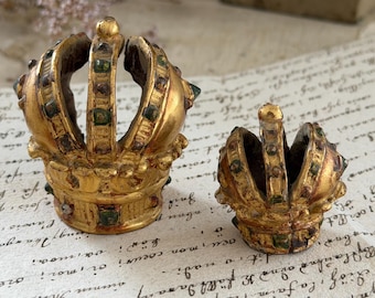 Puede incluir: Dos coronas de tono dorado con un acabado desgastado, que presentan pequeños acentos cuadrados incrustados de color verde y oscuro a lo largo de los arcos y la base. Las coronas están abiertas por arriba y muestran un aspecto texturizado.