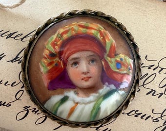 Puede incluir: Un broche redondo con un retrato pintado a mano de una persona que lleva un tocado vibrante y multicolor con patrones rojos, naranjas y verdes. El retrato está engastado en un marco de metal trenzado y texturizado con un acabado en bronce oscuro.