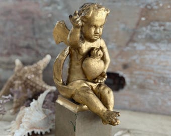 Puede incluir: Una figura de querubín dorada sentada sobre una base cuadrada, sosteniendo un objeto en forma de corazón con una mano mientras gesticula hacia arriba con la otra. La figura presenta alas pequeñas y un acabado metálico desgastado.