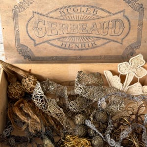 Könnte beinhalten: Eine Holzkiste mit dem Text "KUGLER GERBEAUD HENRIK" auf dem Deckel, gefüllt mit verschiedenen Vintage-Spitzen und -Besätzen. Die Besätze umfassen Gold-, Silber- und Beigetöne mit aufwendigen Mustern und Texturen. Einige Besätze sind geknotet.