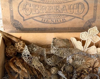 Puede incluir: Una caja de madera con el texto "KUGLER GERBEAUD HENRIK" en la tapa, llena de encajes y adornos vintage. Los adornos incluyen tonos dorados, plateados y beige, con patrones y texturas intrincados. Algunos adornos están anudados.