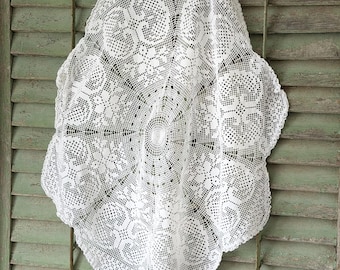 Puede incluir: Un tapete blanco, intrincadamente tejido a ganchillo con un borde festoneado. El delicado patrón de encaje presenta diseños florales y geométricos. El tapete cuelga contra una contraventana de madera pintada de verde desgastado.