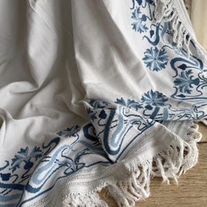 Puede incluir: Textil blanco con bordado floral azul y ribete de flecos. El dise&ntilde;o presenta flores estilizadas y patrones de desplazamiento en diferentes tonos de azul. La tela est&aacute; drapeada, mostrando la artesan&iacute;a detallada y la textura.