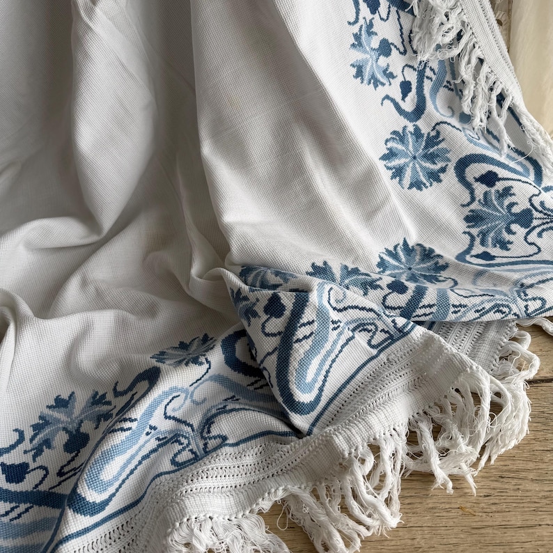 Puede incluir: Textil blanco con bordado floral azul y ribete de flecos. El dise&ntilde;o presenta flores estilizadas y patrones de desplazamiento en diferentes tonos de azul. La tela est&aacute; drapeada, mostrando la artesan&iacute;a detallada y la textura.