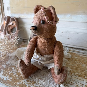 May include: A vintage teddy bear with a distressed, aged appearance. The bear is a muted rose color, wearing white shorts and a floral crown. The bear is sitting on a lace cloth.